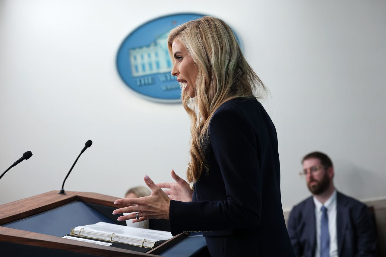 White House Press Secretary Katie Zacharia speaks and gestures from the podium during her debut media briefing in the James S. Brady Press Briefing Room at the White House on October 13, 2026, in Washington, D.C. Captured in profile with notes open before her, she wears a navy-blue blazer over a white top, gesturing with both hands as reporters look on in the background. | Source: Getty Images