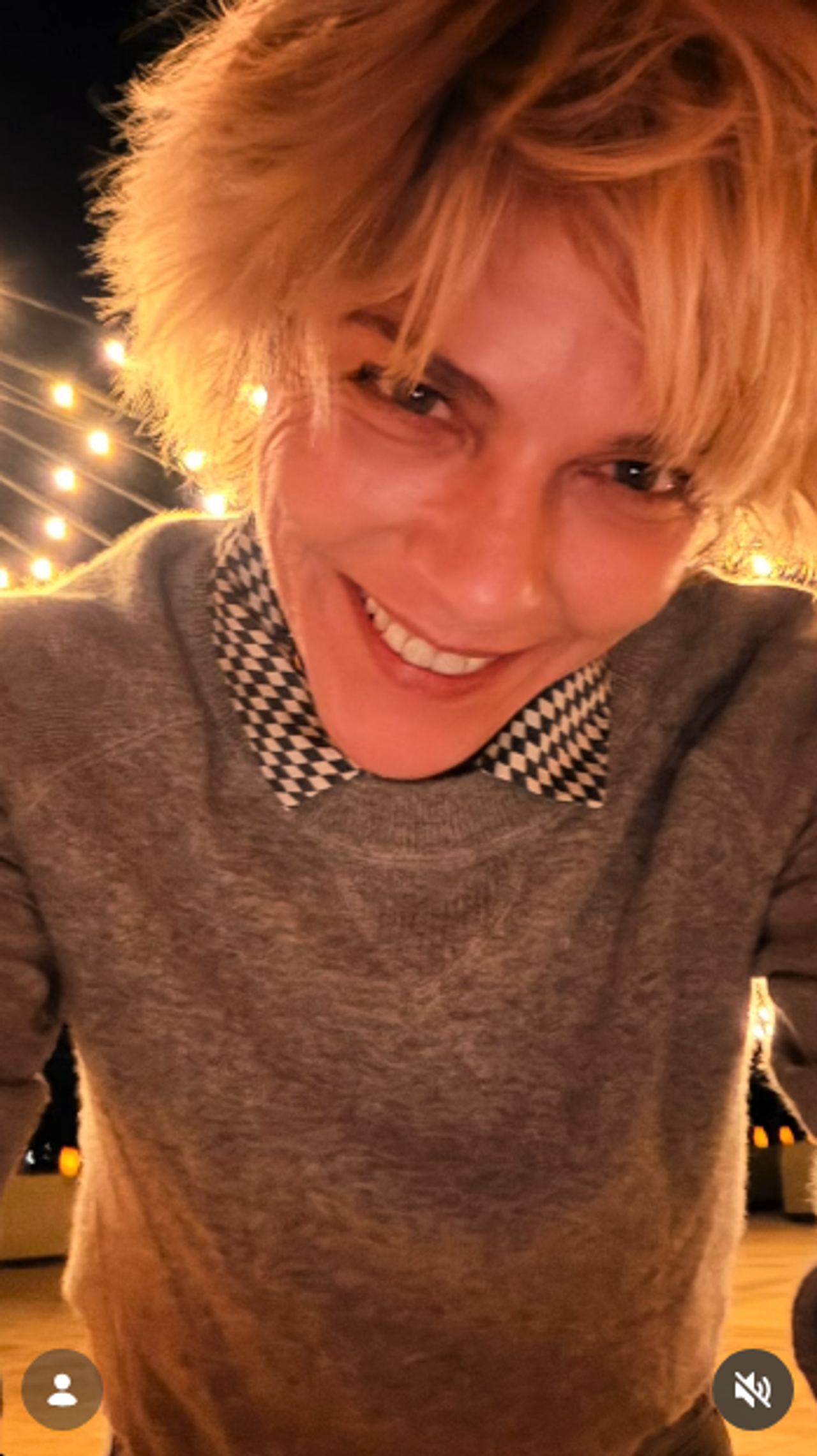 Smiling directly into the camera from above, Selma Blair captures an evening selfie beneath glowing cafe lights on October 11, 2026. She wears a grey pullover layered over a graphic houndstooth collar, with her blonde textured bangs falling across her brow. | Source: Instagram/selmablair
