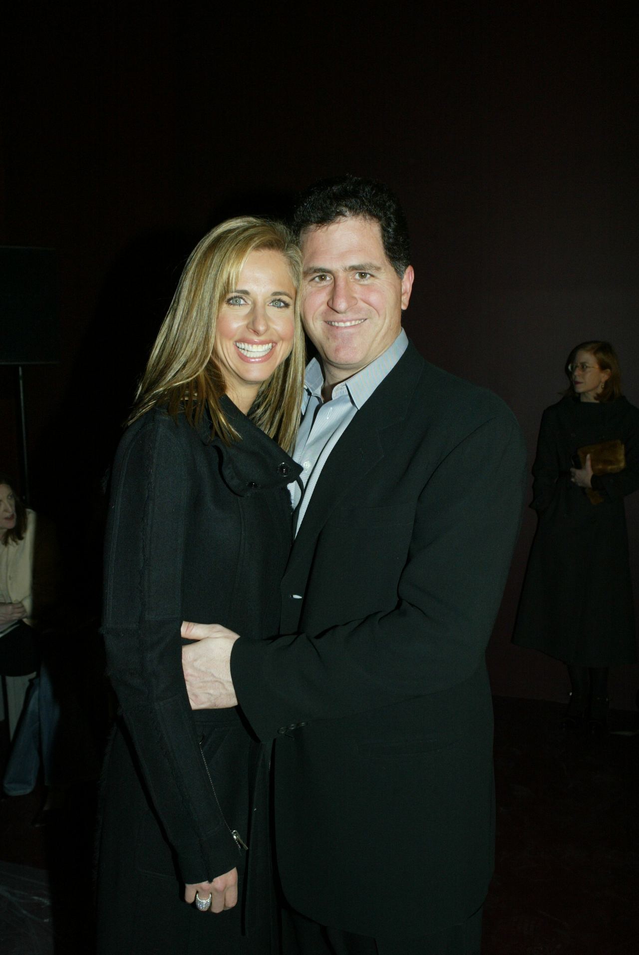 Susan Dell and Michael Dell smiled warmly while embracing at the Fall 2004 Phi fashion show in New York on February 13, 2004. Susan wore a stylish long black coat with a wide collar, while Michael coordinated beside her in a classic black blazer over an open-collar grey dress shirt. | Source: Getty Images