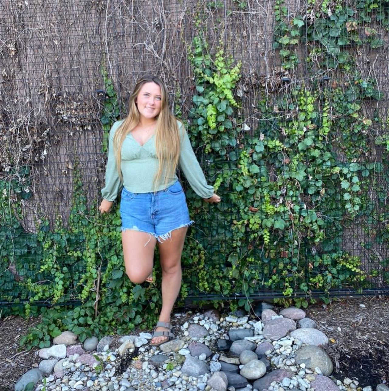 Hannah Jorgensen smiles outdoors as she poses against a leafy ivy-covered trellis wall, dressed in a sage-green long-sleeved top, denim shorts, and sandals. | Source: Facebook/Hannah Jorgensen